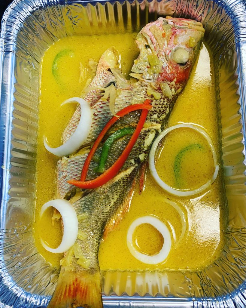 A fish resting in a tin container surrounded by sliced onions and colorful peppers.